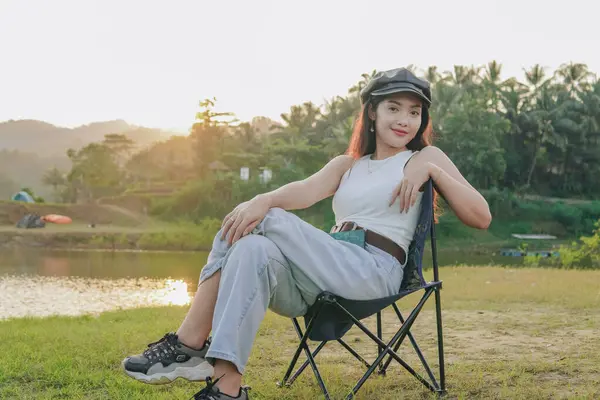 stock image Joyful young Asian woman is sitting in a lawn chair by a lake while smiling to the camera, nature background.