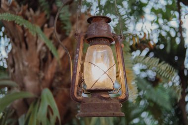 İçinde ışık olan bir ağaçtan sarkan bir gaz lambası. Fener eski ve paslı. Sahnenin sıcak ve rahat bir atmosferi var..