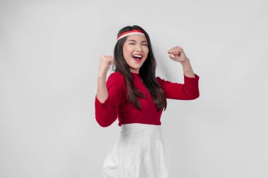 Kırmızı tişörtlü ve beyaz pantolonlu aşırı neşeli genç Asyalı kadın mutlu ve başarılı bir ifadeyle ellerini havaya kaldırıyor. Endonezya Bağımsızlık Günü kavramı.