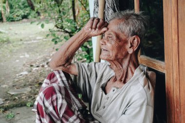 Thoughtful senior elderly man wearing saroong sitting alone in front of house with hands placed on head.