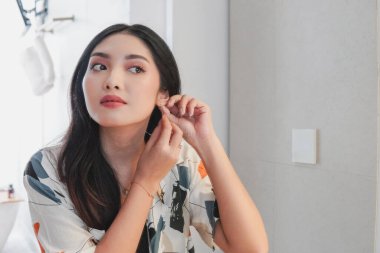 Elegant Asian woman is getting ready to go out and is putting on her earrings. She is wearing a white shirt with a colored pattern.