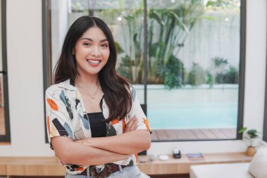 Confident Asian woman is smiling and posing for a picture with a folded arms in front of a pool. Concept of relaxation and leisure.