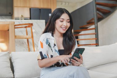 Excited young Asian woman is sitting on a couch and looking at her cell phone with villa kitchen island in the background.