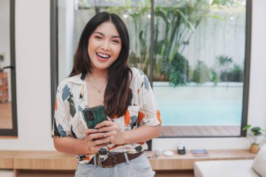 Joyful young Asian woman is smiling and holding smartphone, posing in a room with swimming pool outside.