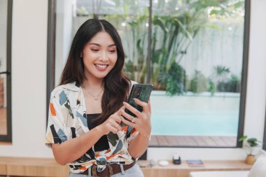 Joyful young Asian woman is smiling and holding smartphone, posing in a room with swimming pool outside.