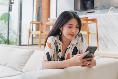 Huzurlu genç Asyalı kadın bir kanepede oturuyor ve cep telefonuna bakıyor. Gülümsüyor ve sosyal medyada boş zamanların tadını çıkarıyor..