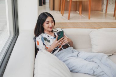 Huzurlu genç Asyalı kadın bir kanepede oturuyor ve cep telefonuna bakıyor. Gülümsüyor ve sosyal medyada boş zamanların tadını çıkarıyor..