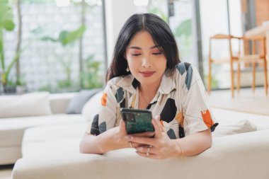 Huzurlu genç Asyalı kadın bir kanepede oturuyor ve cep telefonuna bakıyor. Gülümsüyor ve sosyal medyada boş zamanların tadını çıkarıyor..