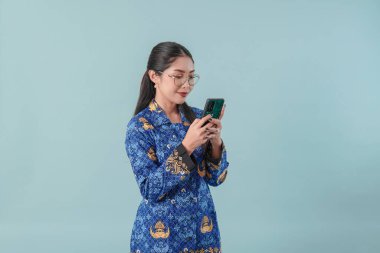 Gülümseyen Endonezyalı devlet çalışanı kadın mavi batik üniforma ve gözlük takıyor interneti taramak için akıllı telefon kullanıyor. PNS devlet memuru kavramı.