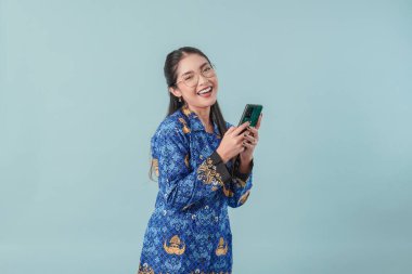 Gülümseyen Endonezyalı devlet çalışanı kadın mavi batik üniforma ve gözlük takıyor interneti taramak için akıllı telefon kullanıyor. PNS devlet memuru kavramı.