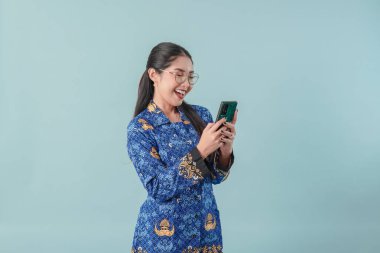 Gülümseyen Endonezyalı devlet çalışanı kadın mavi batik üniforma ve gözlük takıyor interneti taramak için akıllı telefon kullanıyor. PNS devlet memuru kavramı.