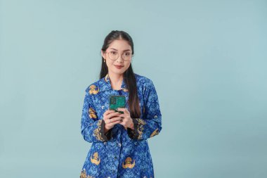 Gülümseyen Endonezyalı devlet çalışanı kadın mavi batik üniforma ve gözlük takıyor interneti taramak için akıllı telefon kullanıyor. PNS devlet memuru kavramı.