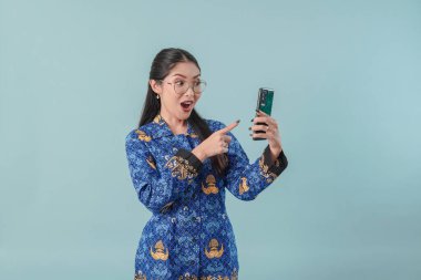 Endonezyalı devlet çalışanı mavi batik üniformalı ve gözlüklü kadın elinde tuttuğu cep telefonunu şaşırmış bir şekilde gösteriyor. PNS devlet memuru kavramı.