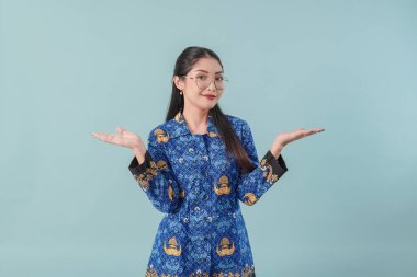 Confused government worker woman wearing blue batik uniform and glasses pointing to the copy space beside her, choosing option. PNS concept. clipart