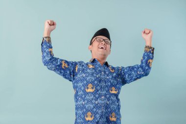 Happy excited government worker man wearing blue batik uniform raising hands freedom sign. PNS civil servant concept. clipart