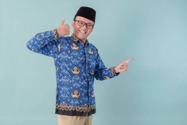 Cheerful government worker man wearing blue batik uniform pointing to the copy space beside him and giving thumb up gesture. PNS civil servant concept. clipart