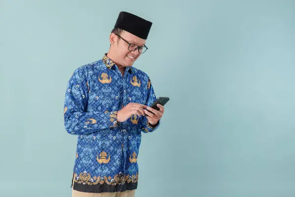 stock image Handsome government worker man wearing blue batik uniform holding smartphone while smiling. PNS and civil servant concept.