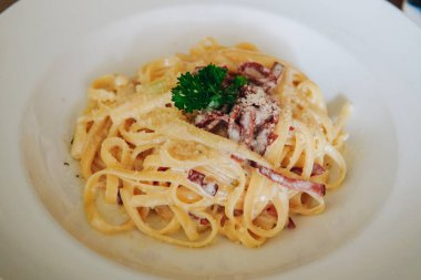 Bir tabak Carbonara fettuccine makarnası. Üstünde yeşil bir bitki var. Tahta masanın üstüne konmuş beyaz bir tabakta servis ediliyor..