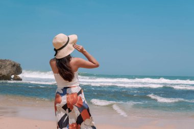 Back portrait of a young Indonesian woman wearing a straw hat enjoying the view of Drini beach in Yogyakarta. Holiday and travel concept.