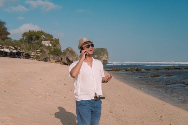 Handsome young Asian man holding his smartphone having a call while walking on the Drini beach, Yogyakarta.