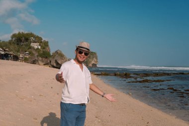 Smiling young Asian man wearing a straw hat and sunglasses doing invite hand gesture while standing by the Drini beach, Yogyakarta.