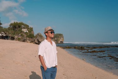 Happy young Asian man wearing a straw hat and sunglasses standing while enjoying scenery on the Drini beach, Yogyakarta.