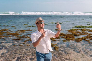 Cheerful young Asian man doing video call using his smartphone while pointing to the scenery of Drini beach, Yogyakarta.