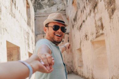 Cheerful young Asian man wearing a straw hat and sunglasses holding hands and pointing to the building of Tamansari Yogyakarta.