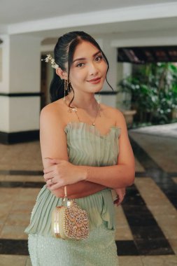 Elegant young Asian woman in a green dress and a flowery accessory is posing for a photo with a smile expression.
