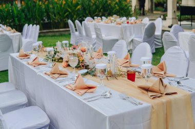 Beyaz masa örtüsü ve ortasında zarif bir çiçek aranjmanı olan uzun masa, bir düğün ya da özel bir etkinlik için hazırlanır..