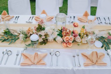 Beyaz masa örtüsü ve ortasında zarif bir çiçek aranjmanı olan uzun bir masa, bir düğün ya da özel bir akşam yemeği için hazırlanır..