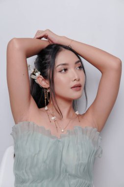 Chic young Asian woman wearing luxurious dress and full make up posing with her arms raised, isolated by white background.