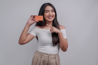 Cheerful young Asian woman pointing to the credit card she holds, isolated by white background.