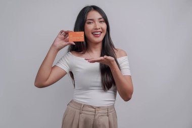 Cheerful young Asian woman pointing to the credit card she holds, isolated by white background.