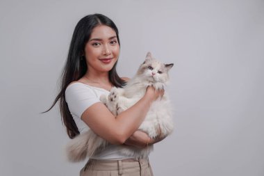 Smiling young Asian woman in white shirt is holding a white ragdoll cat over isolated white background.