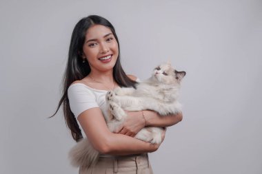 Smiling young Asian woman in white shirt is holding a white ragdoll cat over isolated white background.