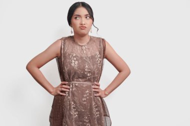 Sad young Asian woman in brown brocade dress standing and placing her arms on hip with a displeased expression, isolated by white background.