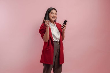 Kırmızı ceketli, gülümseyen ve elinde akıllı telefon tutan güzel bir Asyalı kadın. Arkasındaki fotokopi alanını işaret ediyor. İzole edilmiş pembe arka plan..