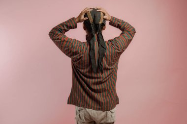 Lurik ve Blangkon giyen Endonezyalı genç bir adamın arka portresi. Kafasını tutuyor. Pembe arka planda izole edilmiş stres ve baş ağrısı hissediyor..