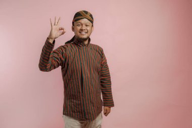 Lurik ve Blangkon giyen mutlu Endonezyalı genç adam pembe arka planla izole edilmiş parmak hareketi yapıyor..