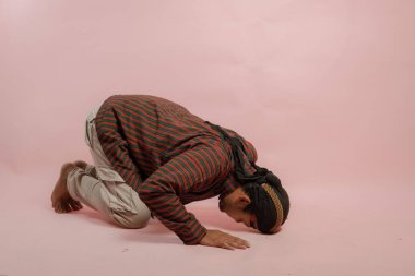Lurik ve blangkon giyen Endonezyalı genç adam, pembe arka planla izole edilmiş, elleri ve başı yerde diz çöküyor..