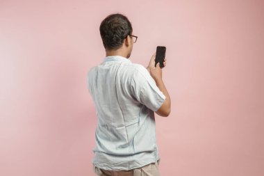 Mavi gömlekli, akıllı telefon kullanan, pembe arka planda yaşayan Asyalı genç bir adamın arka portresi..