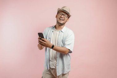 Şapka ve mavi gömlek giyen mutlu genç Asyalı adam, pembe arka planla izole edilmiş akıllı telefonunu tutarken gülüyor.