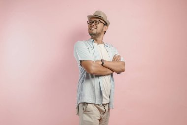 Mavi gömlekli ve şapkalı, pembe arka planda katlanmış kollarıyla poz veren kendine güvenen Asyalı adam..