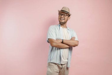 Mavi gömlekli ve şapkalı, pembe arka planda katlanmış kollarıyla poz veren kendine güvenen Asyalı adam..