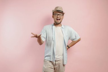 Mavi tişörtlü ve şapkalı neşeli genç Endonezyalı adam pembe arka planla izole edilmiş davetkar bir jest yapıyor..