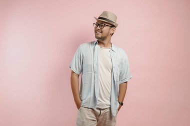 Şapkalı ve mavi gömlekli yakışıklı genç bir adam. Gülümseyerek poz veriyor. Pembe arka planda soyutlanmış..