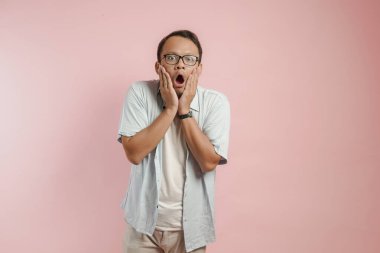 Şok olmuş bir ifadeyle çenesini tutan gözlüklü, pembe arka planlı, şaşırmış genç Asyalı adam..