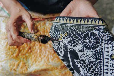 Person's hand is tracing design the pattern of batik tulis cloth using Canting to draw a malam in white cloth.