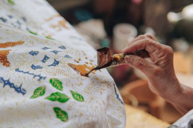 Close up picture of a person's hand drawing the design pattern of Indonesian batik tulis fabric using Canting and malam wax.